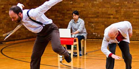 Two men jumping and bowing, while a woman sits behind them using a laptop.